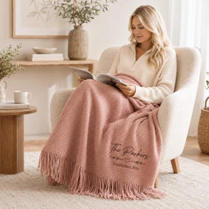 Puede incluir: Una manta de punto texturizado en color rosa empolvado con flecos, que presenta el texto bordado en negro The Parkers junto a un pequeño icono de corazón y las palabras Established 2026, sobre un sillón tapizado de color crema.