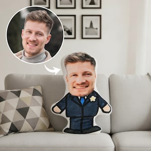 May include: A custom-made pillow shaped like a person, featuring a smiling face and a suit. The pillow is on a gray sofa with a patterned pillow. A photo of the person is in the upper left corner.