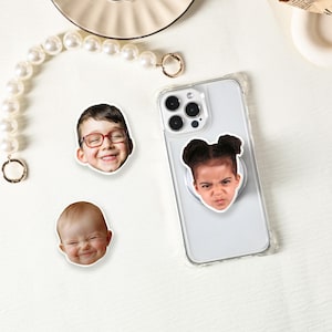 Puede incluir: Una funda transparente para teléfono con un agarre emergente con la cara de un niño. También son visibles dos pegatinas adicionales con caras de niños. Las pegatinas y el agarre emergente tienen un borde blanco. El fondo es blanco.