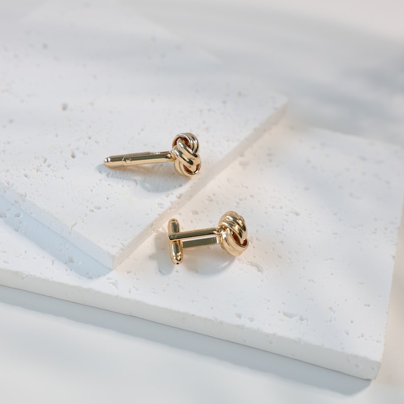 Puede incluir: Dos gemelos dorados con dise&ntilde;o de nudo, presentados sobre una superficie blanca texturizada. El detalle del nudo es una caracter&iacute;stica clave, aportando elegancia. Un accesorio atemporal, adecuado para ocasiones formales.