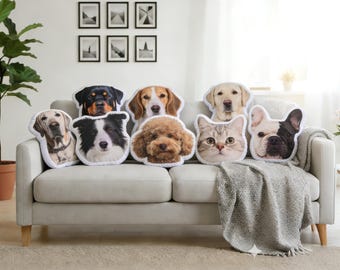 May include: A collection of pet-themed throw pillows featuring various dog and cat breeds. The pillows are arranged on a light gray sofa with a gray throw blanket. The background includes framed artwork and a potted plant.