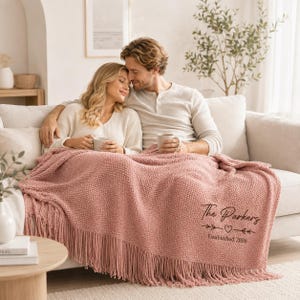 May include: A dusty rose textured throw blanket with fringe trim, featuring black embroidered text that reads The Parkers, a small heart icon, and the words Established 2026. The blanket is draped over a sofa, showcasing its waffle-knit pattern.