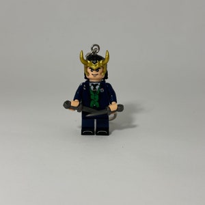 Puede incluir: Una minifigura de Lego de Loki del Universo Cinematográfico de Marvel. La figura lleva un traje azul oscuro con una camisa verde y un casco dorado con cuernos. Sostiene dos dagas plateadas.