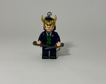 Puede incluir: Una minifigura de Lego de Loki del Universo Cinematográfico de Marvel. La figura lleva un traje azul oscuro con una camisa verde y un casco dorado con cuernos. Sostiene dos dagas plateadas.