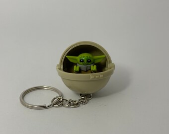 May include: A green and gray keychain featuring a small Lego figure of Baby Yoda in a pod. The keychain is made of plastic and has a silver metal ring.