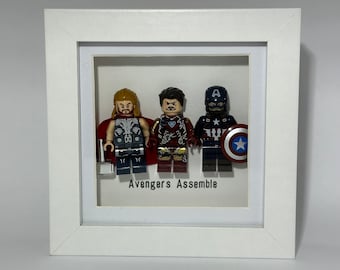 May include: A white frame with a glass front displays three miniature Lego figures of Thor, Iron Man, and Captain America. The figures are arranged in a row, and the frame has the text "Avengers Assemble" printed below the figures.