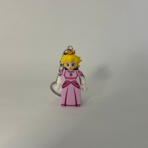 May include: A pink and white Lego-style keychain featuring Princess Peach from the Super Mario franchise. She is wearing a pink dress with a blue gem and a gold crown.