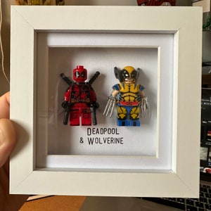 May include: A white frame with a glass front displays two Lego minifigures. The minifigures are Deadpool and Wolverine. The text "DEADPOOL & WOLVERINE" is printed below the figures.