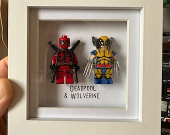 Puede incluir: Un marco blanco con un frente de vidrio muestra dos minifiguras de Lego. Las minifiguras son Deadpool y Wolverine. El texto "DEADPOOL & WOLVERINE" está impreso debajo de las figuras.