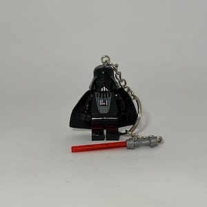 Puede incluir: Un llavero Lego Darth Vader negro con un sable de luz rojo. El llavero tiene una capa negra y una cadena plateada.