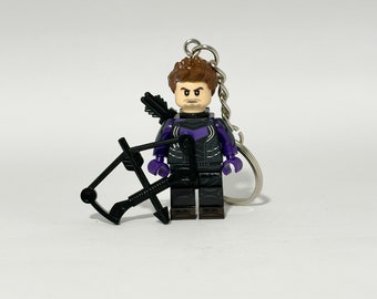 Puede incluir: Una minifigura LEGO de Hawkeye en morado y negro con un arco y una flecha. La minifigura está unida a un llavero plateado.