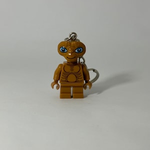 Peut inclure: Une figurine Lego brune du personnage extraterrestre E.T. avec des yeux bleus. La figurine est attachée à un porte-clés argenté.