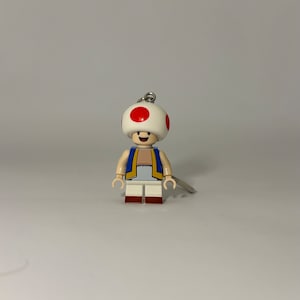 May include: A Lego-style keychain featuring the character Toad from the Super Mario video game series. The figure has a white mushroom-shaped head with red spots, a blue vest, and a smiling face. The keychain is attached with a silver ring.