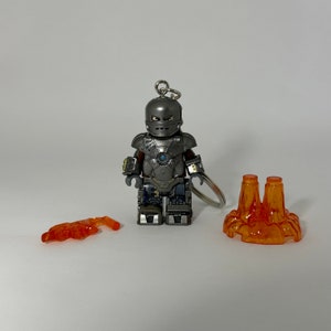 May include: A silver Iron Man Lego minifigure keychain with a red and orange lava base. The minifigure has a blue chest piece and a silver helmet with a blue visor.