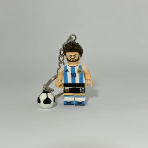 Puede incluir: Una camiseta de fútbol azul y blanca con el número 10 en la parte delantera. La camiseta tiene el logotipo de Adidas y el logotipo de la selección nacional argentina. La figura sostiene un balón de fútbol.