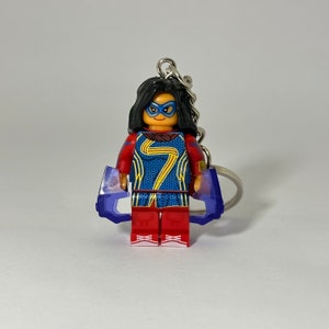 May include: A keychain featuring a Lego minifigure of Captain Marvel, wearing a red and blue costume with a gold symbol on the chest. The figure is holding two purple translucent pieces in each hand. The keychain has a silver ring.
