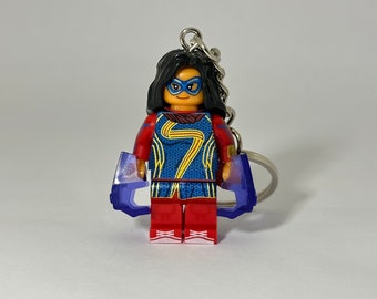 May include: A keychain featuring a Lego minifigure of Captain Marvel, wearing a red and blue costume with a gold symbol on the chest. The figure is holding two purple translucent pieces in each hand. The keychain has a silver ring.