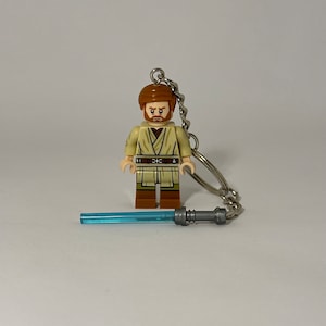 May include: A Lego minifigure of Obi-Wan Kenobi with a brown robe and a blue lightsaber. The minifigure is attached to a silver keychain.