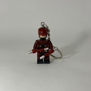 May include: A red and black Lego minifigure of Daredevil holding two silver batons. The minifigure is attached to a silver keychain.