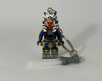 Puede incluir: Un llavero con una figura estilo Lego de Ahsoka Tano de Star Wars. La figura tiene un diseño detallado con detalles en azul, blanco y naranja. Un sable de luz plateado está adjunto. El llavero está en un anillo plateado.