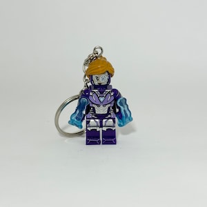 May include: A purple Lego-style keychain figure with a silver and blue design. The figure has a light brown hairstyle and a silver eye. The figure has blue translucent arm attachments. The keychain is attached to a silver ring.