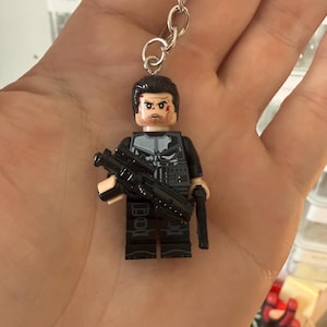 May include: A keyring featuring a plastic minifigure character with dark hair, a beard, and a white skull emblem on a black tactical vest. The figure holds a black toy rifle in its right hand and a black handgun in its left hand, attached to a metal ring.