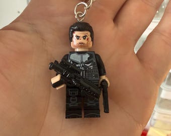 Puede incluir: Un llavero con una minifigura de plástico con cabello oscuro, barba y un emblema de calavera blanca en un chaleco táctico negro. La figura sostiene un rifle de juguete negro en la mano derecha y una pistola negra en la mano izquierda, unido a una argolla.