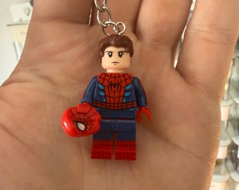 Puede incluir: Un llavero de minifigura de Spider-Man que muestra un personaje con cabello castaño y rostro descubierto. La figura viste un traje azul y rojo con un emblema de araña en el pecho y sostiene una máscara roja de Spider-Man separada en una mano.