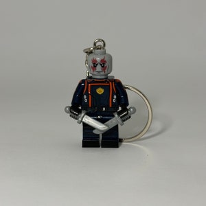 May include: A grey and blue Lego minifigure of a character from Guardians of the Galaxy, holding two silver swords. The minifigure has red markings on its face and is attached to a silver keyring.