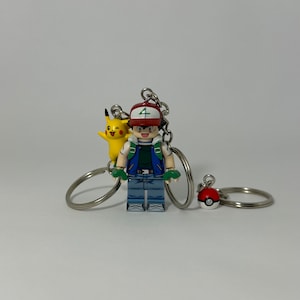 May include: A keychain featuring a Lego-style figure of a Pokemon trainer wearing a red and white hat, a green and blue vest, and blue jeans. The trainer is holding a green and yellow Pokemon figure. The keychain also includes a red and white Pokeball charm.