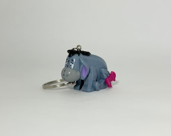 May include: A gray and blue Eeyore keychain with a pink bow. The keychain is made of plastic and has a metal ring.