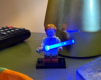 May include: A blue lightsaber is held by a Lego minifigure of a Jedi Knight. The minifigure is wearing a brown robe and has a light brown hairpiece. The lightsaber is lit up and glowing blue.