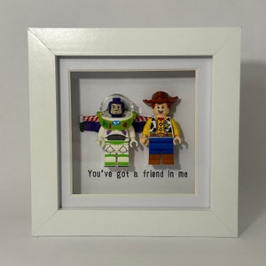 May include: A white frame with a white mat and a glass front. Inside the frame are two Lego minifigures, one of Buzz Lightyear and one of Woody from the Toy Story movies. The quote "You've got a friend in me" is printed below the figures.