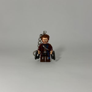 May include: A brown and blue Lego minifigure of Star-Lord from Guardians of the Galaxy, holding two silver blasters. The minifigure is attached to a silver keychain.