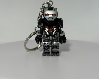 Puede incluir: Un llavero con una figura de acción de War Machine. La figura es una versión en miniatura del personaje, con un casco plateado y ojos rojos. La figura es principalmente negra y plateada, con detalles rojos. El llavero tiene una cadena y un anillo plateados.