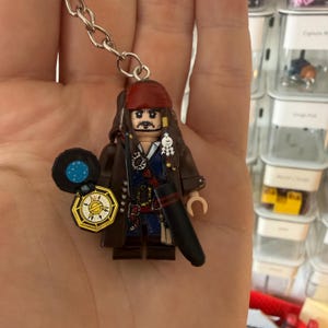 Może przedstawiać: Brelok z plastikową minifigurką pirata w czerwonej chustce na głowie, z długimi brązowymi włosami, w ciemnobrązowym płaszczu i niebieskiej kamizelce. Figurka trzyma mały czarno-złoty kompas z nadrukiem gwiazdy na tarczy.