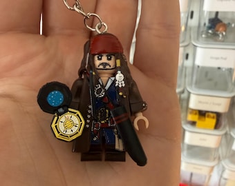 Puede incluir: Un llavero con una minifigura de plástico de un pirata con pañuelo rojo, pelo largo castaño, abrigo marrón oscuro y chaleco azul. La figura sostiene un pequeño accesorio de brújula negro y dorado con un diseño de estrella en la cara.