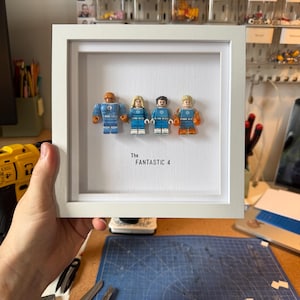 May include: A framed artwork featuring four Lego figures representing the Fantastic Four. The figures are in blue and orange costumes, with the text "The Fantastic 4" below them. The frame is white.