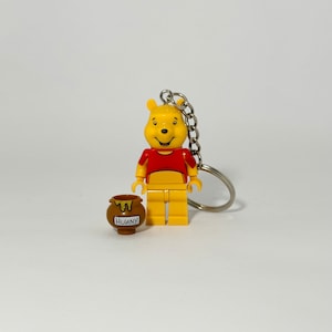 May include: A yellow Winnie the Pooh LEGO minifigure keychain holding a brown honey pot with the word "Hunny" on it. The keychain is attached to a silver metal ring.