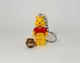 May include: A yellow Winnie the Pooh LEGO minifigure keychain holding a brown honey pot with the word "Hunny" on it. The keychain is attached to a silver metal ring.