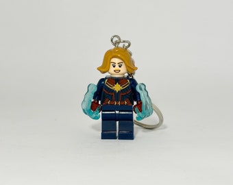May include: A blue and red Lego minifigure of Captain Marvel with blonde hair and a star on her chest. The minifigure is holding blue energy blasts in each hand and is attached to a silver keychain.