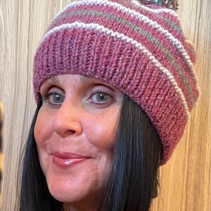 May include: A knitted winter hat in shades of pink, white, and green. The hat features horizontal stripes and a pom-pom on top. The hat is worn by a person with dark hair.