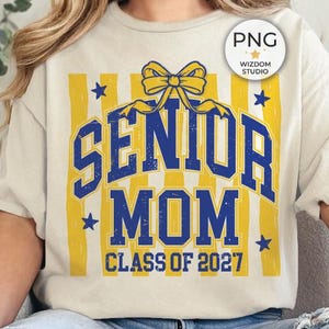 Puede incluir: Una camiseta de color crema con un diseño gráfico de rayas verticales amarillas, un lazo amarillo en la parte superior y texto azul que dice SENIOR MOM CLASS OF 2027, acentuado con pequeñas estrellas azules en el área central de impresión.