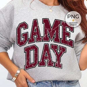 May include: A heather gray sweatshirt with the words "GAME DAY" in a distressed maroon font. The sweatshirt has long sleeves and a crew neck. The text is in a bold, collegiate style. The sweatshirt is paired with blue jeans.
