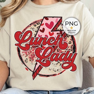 Puede incluir: Una mujer lleva una camiseta beige claro con un gráfico rojo y rosa que dice "Lunch Lady" con un diseño de rayo. El gráfico tiene un estampado de leopardo y corazones.