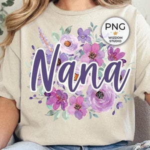 May include: A beige t-shirt with the word "Nana" in purple script, surrounded by watercolor-style purple and lavender flowers. The shirt is part of a casual outfit, paired with blue jeans.