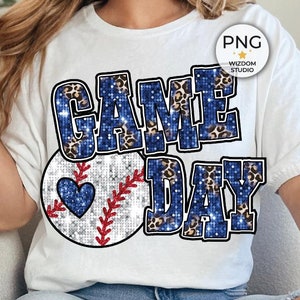 May include: A white t-shirt with the text "GAME DAY" in blue and gold glitter letters with a baseball graphic. The baseball has a blue and gold glitter design with a heart in the center.