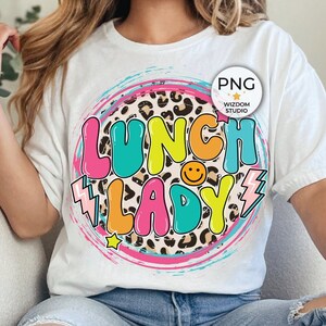 Puede incluir: Una camiseta blanca con un diseño gráfico colorido que dice "Lunch Lady" en un círculo con un fondo de estampado de leopardo. El gráfico también incluye un rayo y una carita sonriente.