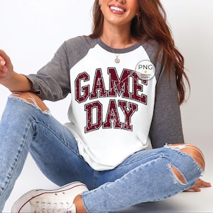 May include: White and gray raglan-style shirt with the text "GAME DAY" in maroon block letters. The shirt has gray sleeves and a white body. The text has a distressed, vintage look.