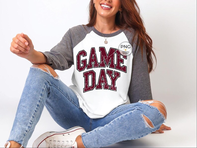 May include: White and gray raglan-style shirt with the text "GAME DAY" in maroon block letters. The shirt has gray sleeves and a white body. The text has a distressed, vintage look.